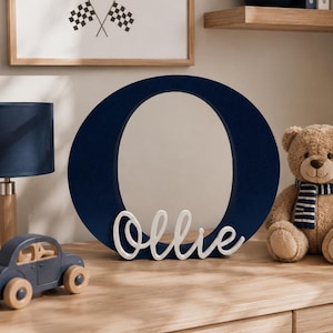 Puede incluir: Una letra "O" azul marino con el nombre "Ollie" en escritura blanca. Tambi&eacute;n se ve una impresi&oacute;n enmarcada de un coche de carreras azul y un oso de peluche con una bufanda a rayas. La decoraci&oacute;n est&aacute; sobre una c&oacute;moda de madera.