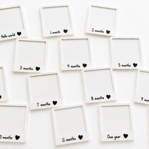 May include: A set of fourteen white square frames shaped like instant photos. Each frame features black printed text and a small heart icon. The text includes Hello world, 1 month, 2 months, 3 months, 4 months, 5 months, 6 months, 7 months, 8 months, 9 months, 10 months, 11 months, One year, and Love you.