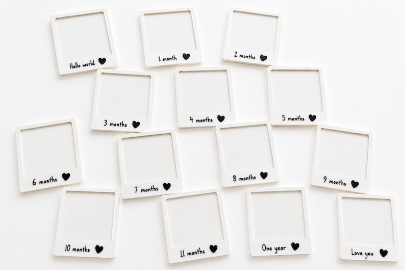 May include: A set of fourteen white square frames shaped like instant photos. Each frame features black printed text and a small heart icon. The text includes Hello world, 1 month, 2 months, 3 months, 4 months, 5 months, 6 months, 7 months, 8 months, 9 months, 10 months, 11 months, One year, and Love you.