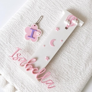 Puede incluir: Un conjunto de decoraci&oacute;n de guarder&iacute;a personalizado con una letra de madera blanca "I" con estrellas rosas, una luna creciente y un globo aerost&aacute;tico. Tambi&eacute;n se incluye un llavero en forma de flor rosa con la letra "I" y el nombre "Isabella" en escritura rosa.