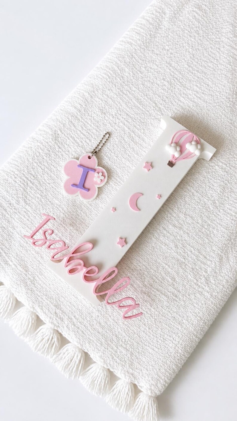 Puede incluir: Un conjunto de decoraci&oacute;n de guarder&iacute;a personalizado con una letra de madera blanca "I" con estrellas rosas, una luna creciente y un globo aerost&aacute;tico. Tambi&eacute;n se incluye un llavero en forma de flor rosa con la letra "I" y el nombre "Isabella" en escritura rosa.