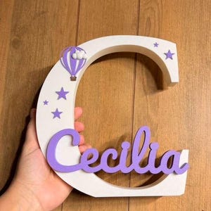 May include: A white wooden letter "C" with the name "Cecilia" in purple script. The letter is decorated with purple stars and a purple hot air balloon. A hand holds the letter against a wooden background.
