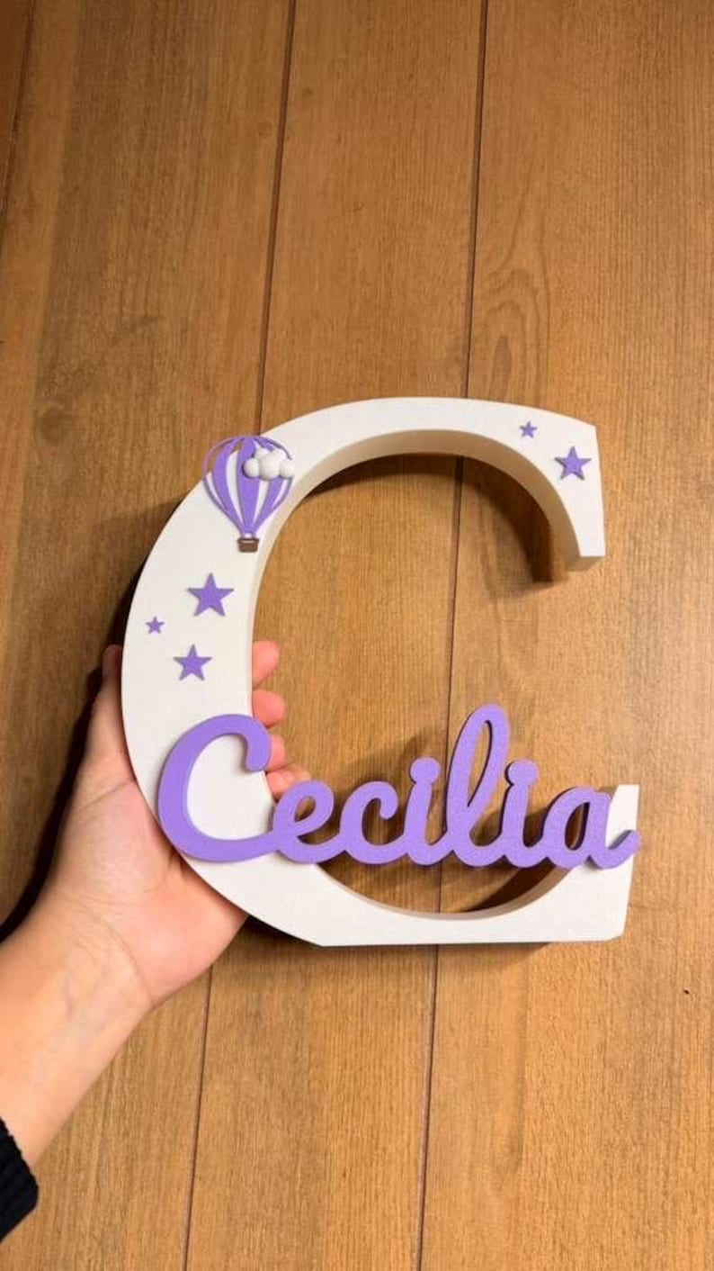 May include: A white wooden letter "C" with the name "Cecilia" in purple script. The letter is decorated with purple stars and a purple hot air balloon. A hand holds the letter against a wooden background.