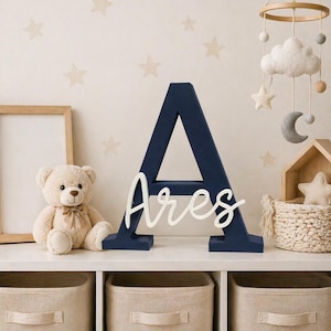 May include: A navy blue wooden letter "A" with the name "Ares" in white script. A beige teddy bear, a framed picture, and a decorative mobile with clouds and stars are on a white shelf. Beige storage bins are below.