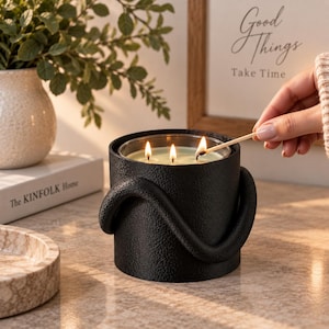Puede incluir: Un portavelas cil&iacute;ndrico negro con una superficie texturizada y un acento curvo similar a un asa. El portavelas contiene una vela de tres mechas con cera de color claro, que est&aacute; siendo encendida con una cerilla de madera.