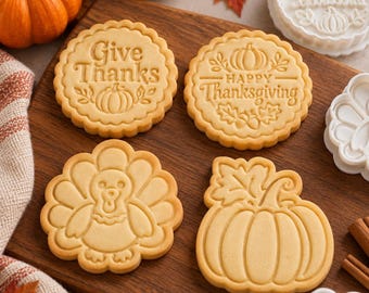 Puede incluir: Cuatro galletas redondas y con formas que presentan diseños en relieve, incluyendo una calabaza, un pavo y el texto Give Thanks y Happy Thanksgiving. Las galletas son de color tostado claro con bordes festoneados y cortadores blancos.