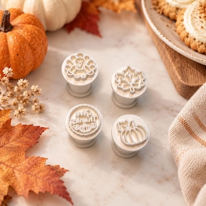 May include: Four white plastic cookie stamps featuring seasonal designs including a turkey, a maple leaf, a pumpkin, and the text "Give Thanks" with a small pumpkin icon. Each stamp has a cylindrical handle for pressing into dough.