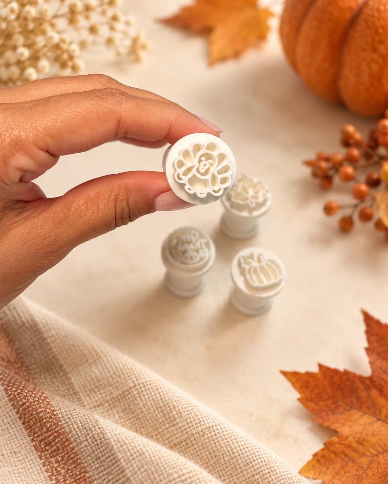 May include: A set of four white plastic plunger-style cookie stamps featuring embossed designs of a turkey, a pumpkin, a maple leaf, and the text Give Thanks. Each stamp has a cylindrical handle and a flat base for pressing dough.