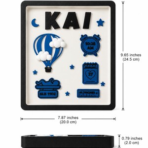 May include: A rectangular black-framed wall sign with a white background featuring the name KAI in black letters. It includes blue icons of a hot air balloon, an alarm clock showing 10:25 AM, a calendar showing February 27, a baby on a scale reading 6LB 11OZ, and a ruler reading 19.7INCHES. Dimensions are 9.65 by 7.87 by 0.79 inches.