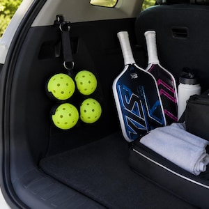 Puede incluir: Un organizador de correa negro colgado de un gancho interior del veh&iacute;culo, que sostiene cuatro pelotas de pickleball amarillas perforadas. Junto a &eacute;l hay dos palas de pickleball con dise&ntilde;os gr&aacute;ficos azules y rosas, una botella de agua y una bolsa.