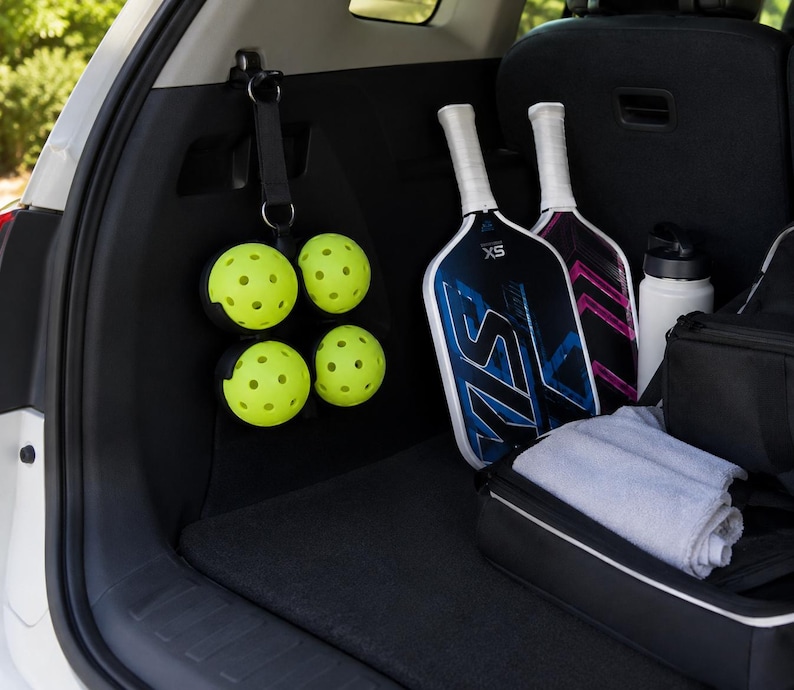 Puede incluir: Un organizador de correa negro colgado de un gancho interior del veh&iacute;culo, que sostiene cuatro pelotas de pickleball amarillas perforadas. Junto a &eacute;l hay dos palas de pickleball con dise&ntilde;os gr&aacute;ficos azules y rosas, una botella de agua y una bolsa.