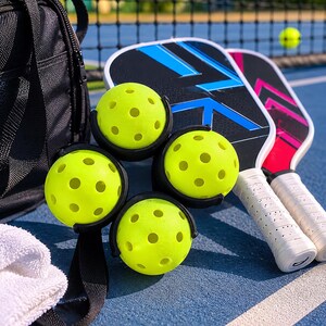 Puede incluir: Cuatro pelotas de pickleball amarillas perforadas sujetas con clips negros, dos palas de pickleball con patrones geom&eacute;tricos azules y rosas, una bolsa de equipo negra y una toalla blanca sobre una pista azul con una red al fondo.