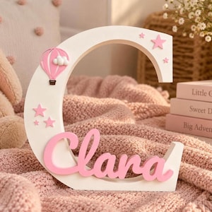 May include: A white wooden letter C featuring the name Clara in pink script lettering. The letter is decorated with a pink hot air balloon, white clouds, and several pink stars of varying sizes attached to the front surface of the letter.