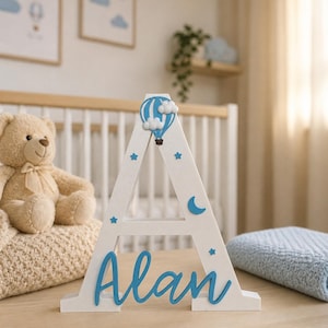 May include: A white wooden letter "A" with the name "Alan" in blue script. The letter is decorated with blue stars, a crescent moon, and a hot air balloon. A teddy bear and a blue blanket are in the background.