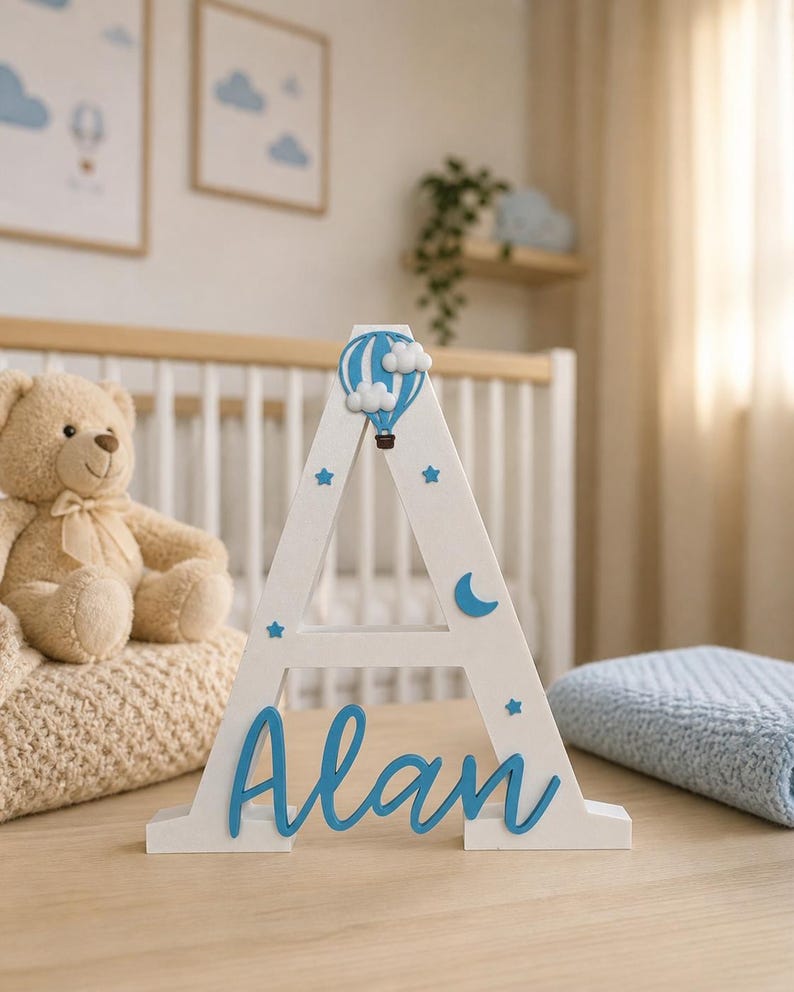 May include: A white wooden letter "A" with the name "Alan" in blue script. The letter is decorated with blue stars, a crescent moon, and a hot air balloon. A teddy bear and a blue blanket are in the background.