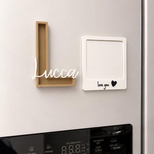 May include: A gold-colored letter L frame with the white script name Lucca attached, and a white square frame featuring the black text love you followed by a black heart symbol, both mounted on a refrigerator surface above the digital control panel.