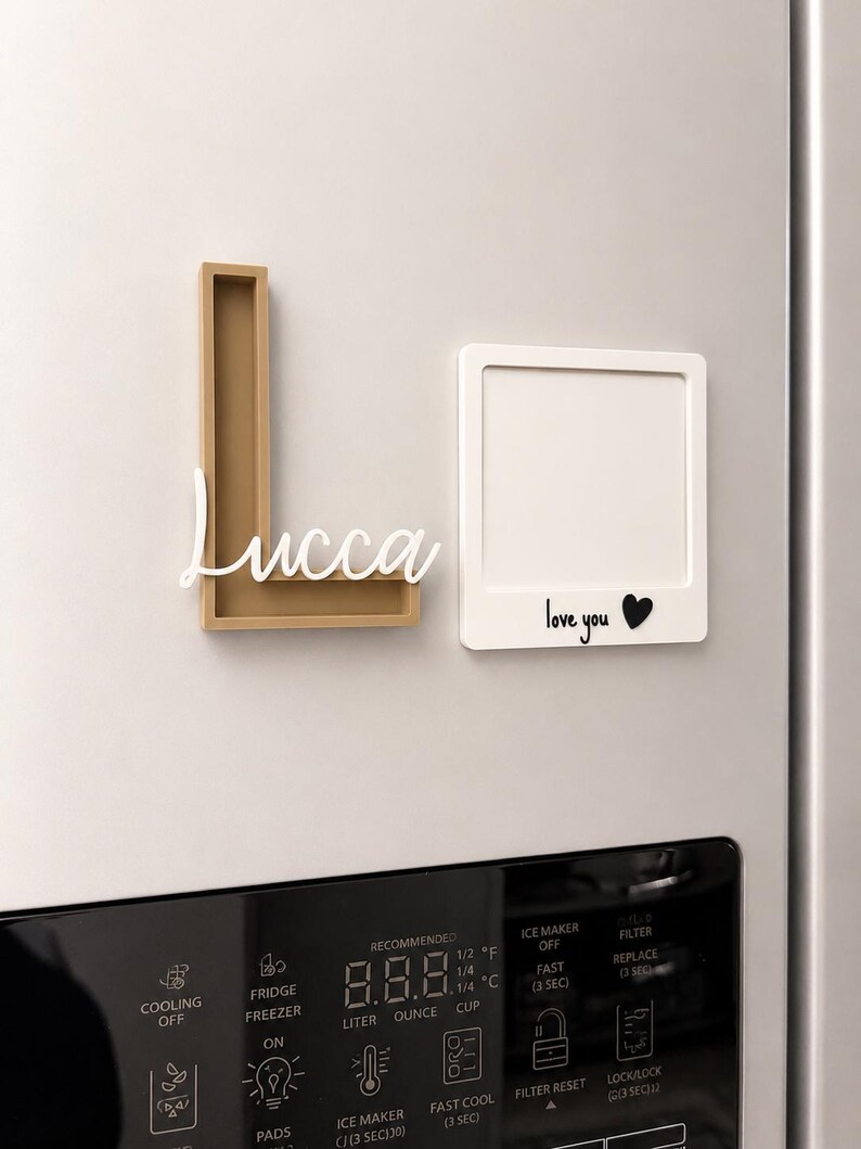 May include: A gold-colored letter L frame with the white script name Lucca attached, and a white square frame featuring the black text love you followed by a black heart symbol, both mounted on a refrigerator surface above the digital control panel.