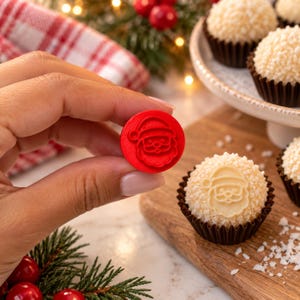 May include: A small, circular red plastic stamp featuring an embossed Santa Claus face design. The stamp is held up to show the detail of the Santa icon, which matches the imprint visible on the white, textured round confection sitting nearby.