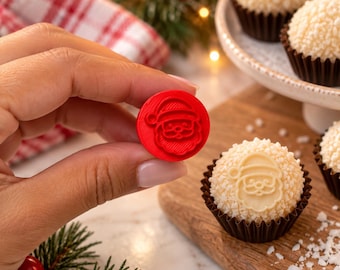 Puede incluir: Un pequeño sello circular de plástico rojo con un diseño en relieve de la cara de Santa Claus. El sello se sostiene para mostrar el detalle del icono de Santa, que coincide con la impresión visible en el dulce redondo y texturizado.