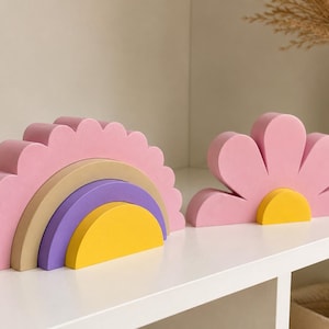 K&ouml;nnte beinhalten: Ein rosa Holz-Regenbogen-Stapelspielzeug mit gewelltem Rand, mit Schichten aus Beige, Lila und Gelb. Daneben ein rosa bl&uuml;tenf&ouml;rmiges Spielzeug mit gelber Mitte. Diese Spielzeuge sind f&uuml;r Kinder.