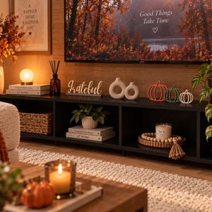 May include: A black media console featuring a white Grateful sign, two beige ceramic donut vases, and three wire pumpkin silhouettes in orange, green, and white. The shelf holds books, a candle on a wooden bead tray, and a woven basket.