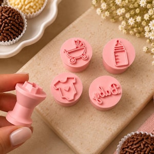 May include: A set of five light pink cylindrical stamps for decorating food. Four stamps feature embossed designs including a baby carriage, a baby bottle, a onesie with a heart, and the word Baby in cursive script. One stamp is held upright.