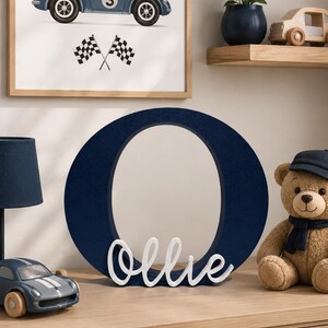 Puede incluir: Una impresi&oacute;n art&iacute;stica enmarcada en azul marino de un coche de carreras vintage con el n&uacute;mero 3. Una gran letra "O" azul marino con el nombre "Ollie" en escritura blanca est&aacute; sobre una c&oacute;moda de madera. Un oso de peluche y un coche de juguete tambi&eacute;n est&aacute;n en la c&oacute;moda.