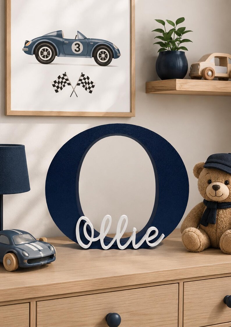 Puede incluir: Una impresi&oacute;n art&iacute;stica enmarcada en azul marino de un coche de carreras vintage con el n&uacute;mero 3. Una gran letra "O" azul marino con el nombre "Ollie" en escritura blanca est&aacute; sobre una c&oacute;moda de madera. Un oso de peluche y un coche de juguete tambi&eacute;n est&aacute;n en la c&oacute;moda.