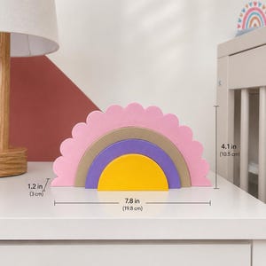 K&ouml;nnte beinhalten: Ein Holz-Regenbogen-Stapelturm in Rosa, Gold, Lila und Gelb. Das Spielzeug ist 19,8 cm breit und 10,5 cm hoch. Die oberste Schicht hat einen gewellten Rand. Ein dekorativer Artikel f&uuml;r ein Kinderzimmer oder Spielzimmer.
