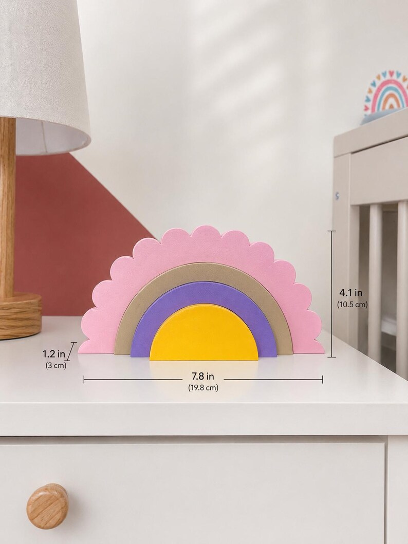 K&ouml;nnte beinhalten: Ein Holz-Regenbogen-Stapelturm in Rosa, Gold, Lila und Gelb. Das Spielzeug ist 19,8 cm breit und 10,5 cm hoch. Die oberste Schicht hat einen gewellten Rand. Ein dekorativer Artikel f&uuml;r ein Kinderzimmer oder Spielzimmer.