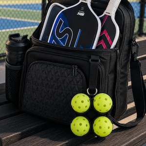 Puede incluir: Una mochila de pickleball negra con un bolsillo frontal texturizado, un portabotellas de malla lateral y dos palas SLK guardadas dentro. Un clip negro sujeta cuatro pelotas de pickleball amarillas perforadas en el exterior.