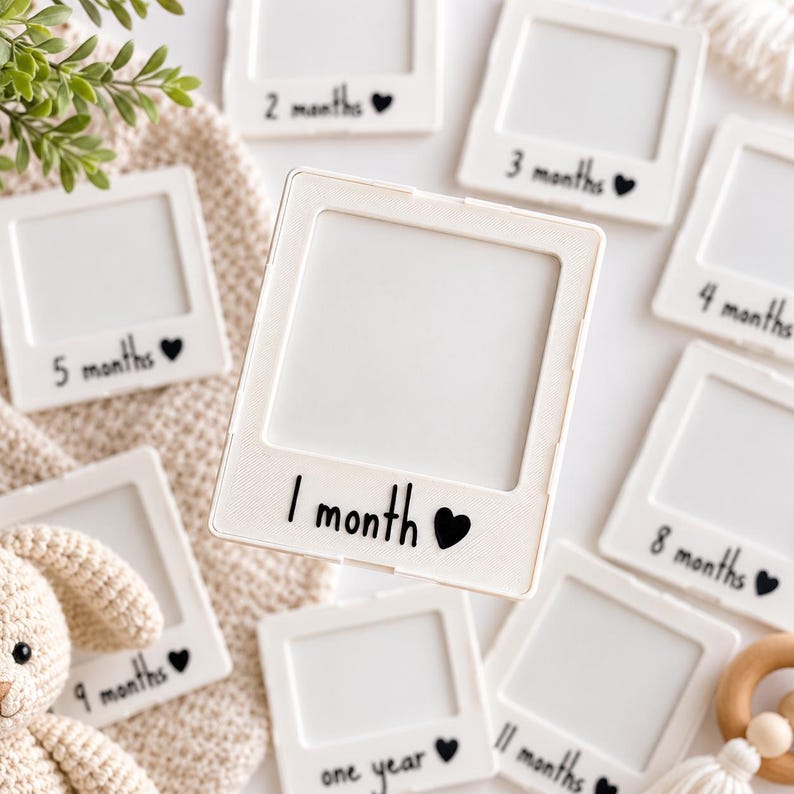 May include: A collection of white square photo frames designed to look like instant film prints. Each frame features black text indicating a baby's age from 1 month to one year, accompanied by a small black heart icon printed below the photo area.