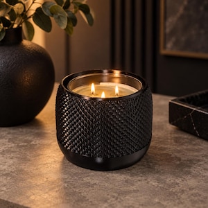 May include: A three-wick candle in a black glass container with a textured, diamond-patterned exterior. The candle is lit, showing three small flames glowing within the clear glass insert. The base of the container is smooth and rounded.