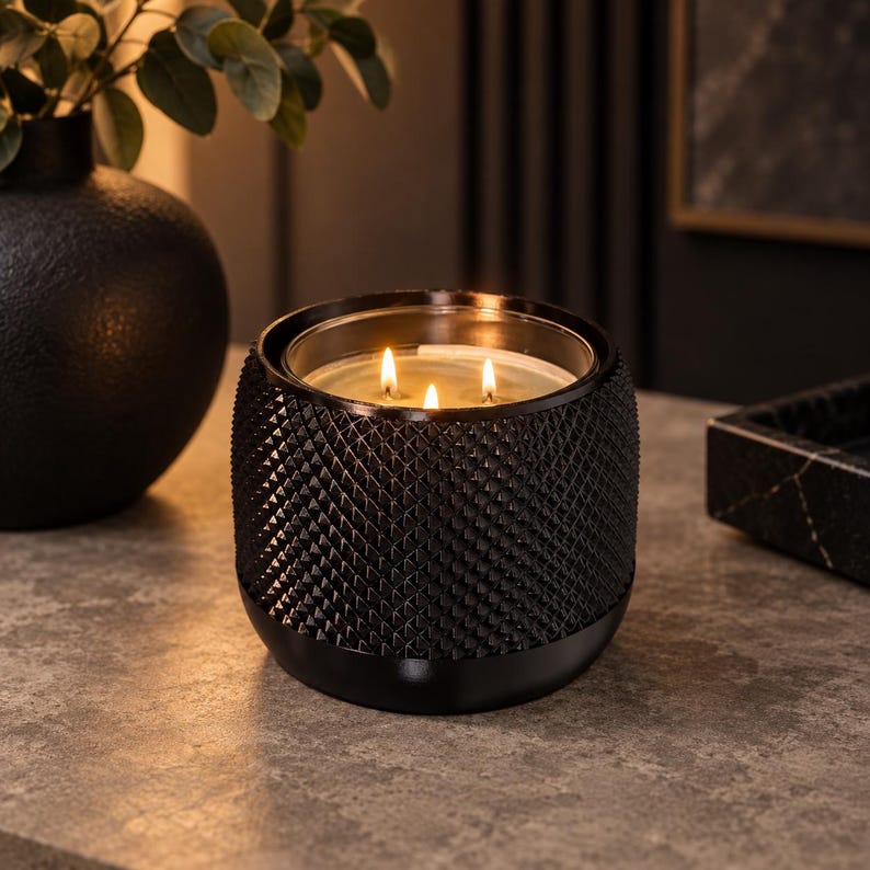 May include: A three-wick candle in a black glass container with a textured, diamond-patterned exterior. The candle is lit, showing three small flames glowing within the clear glass insert. The base of the container is smooth and rounded.