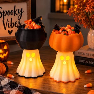 May include: Two illuminated ghost-shaped stands holding candy bowls. One stand supports a black cauldron-shaped bowl, and the other supports an orange pumpkin-shaped bowl. Both ghost bases are white with cut-out eyes and mouths, glowing from within.