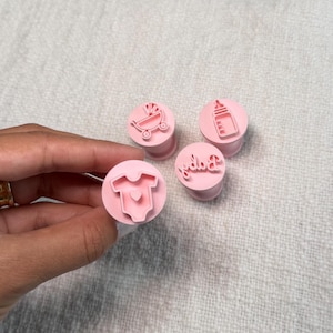 May include: A set of four light pink circular stamps featuring raised designs of a baby onesie with a heart, a baby carriage, a baby bottle, and the word Baby in cursive script. Each stamp has a cylindrical base for easy handling and pressing.