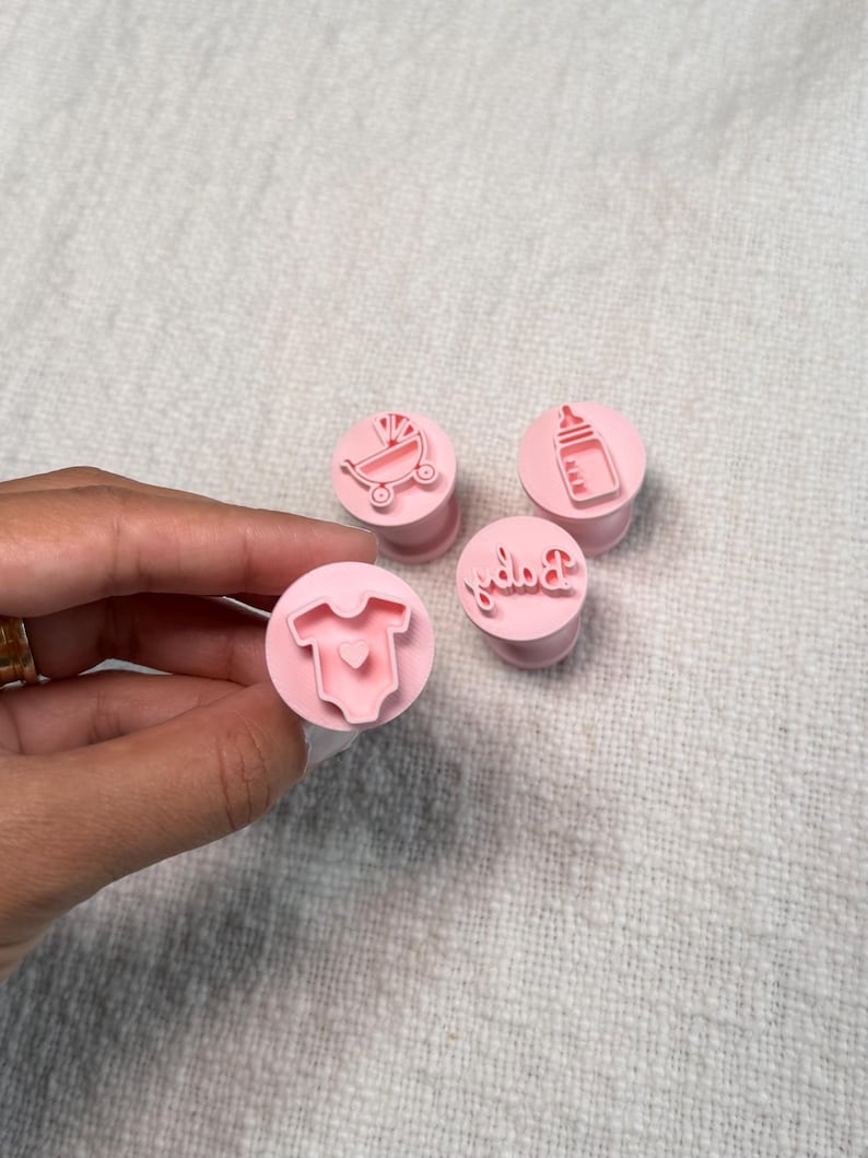 May include: A set of four light pink circular stamps featuring raised designs of a baby onesie with a heart, a baby carriage, a baby bottle, and the word Baby in cursive script. Each stamp has a cylindrical base for easy handling and pressing.