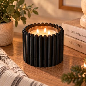 May include: A three-wick candle in a clear glass container, encased in a matte black cylindrical holder with a vertical ribbed, fluted texture. The candle features three lit wicks producing a warm glow, set within the dark, structured exterior.