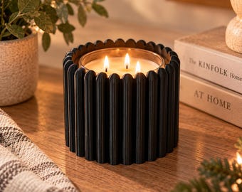 May include: A three-wick candle in a clear glass container, encased in a matte black cylindrical holder with a vertical ribbed, fluted texture. The candle features three lit wicks producing a warm glow, set within the dark, structured exterior.