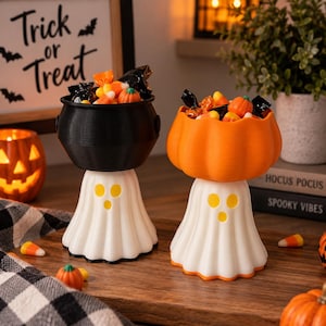 May include: Two decorative candy bowls featuring white ghost-shaped bases with yellow circular eyes and mouths. One bowl is a black cauldron shape, and the other is an orange pumpkin shape. Both are filled with assorted Halloween sweets.