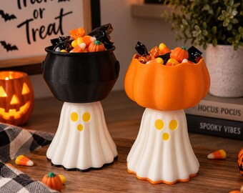 May include: Two decorative candy bowls featuring white ghost-shaped bases with yellow circular eyes and mouths. One bowl is a black cauldron shape, and the other is an orange pumpkin shape. Both are filled with assorted Halloween candies.