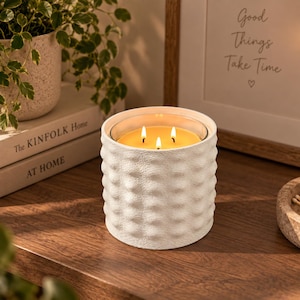 May include: A three-wick candle in a white cylindrical ceramic vessel with a textured, dimpled exterior surface. The candle wax is yellow and illuminated by three small, bright flames, casting a warm glow from within the container.
