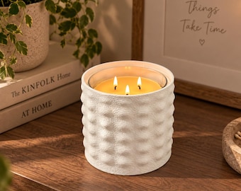 Puede incluir: Una vela de tres mechas en un recipiente cilíndrico de cerámica blanca con una superficie exterior texturizada y con hoyuelos. La cera es amarilla e iluminada por tres pequeñas llamas brillantes que emiten un cálido resplandor.