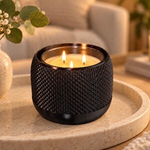 May include: A three-wick candle in a black cylindrical container with a textured, diamond-patterned exterior. The wax is cream-colored, and the three flames are lit, casting a warm glow from within the glass vessel, which has a smooth base.