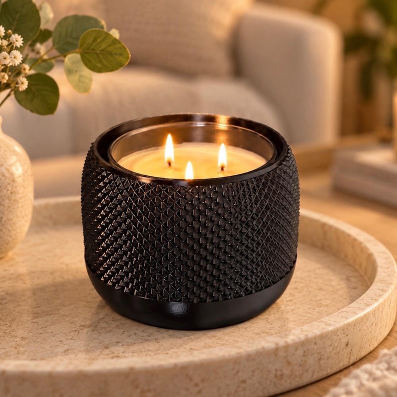 May include: A three-wick candle in a black cylindrical container with a textured, diamond-patterned exterior. The wax is cream-colored, and the three flames are lit, casting a warm glow from within the glass vessel, which has a smooth base.