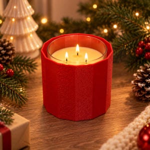May include: A three-wick candle in a clear glass container, encased in a bright red, faceted, textured sleeve. The candle wax is off-white and the three wicks are lit, producing a warm, glowing flame. The exterior of the sleeve has vertical ridges.