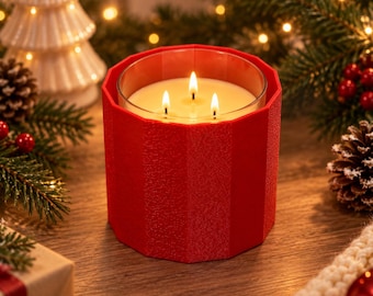 Puede incluir: Una vela de tres mechas en un recipiente de vidrio transparente, dentro de una funda roja brillante, facetada y texturizada. La cera es de color blanco roto y las tres mechas están encendidas, produciendo una llama cálida. Tiene estrías.