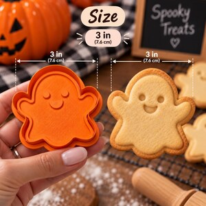 May include: An orange ghost-shaped cookie cutter measuring 3 inches in width, shown alongside a baked ghost-shaped cookie. The cutter features a detailed impression design for the eyes and mouth, matching the baked cookie's appearance.