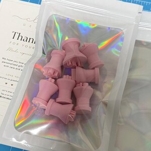 May include: A collection of small, light pink, spool-shaped plastic stamps with flat circular bases and textured tops, contained within a clear, iridescent, resealable plastic pouch that displays a rainbow-like sheen under the light.