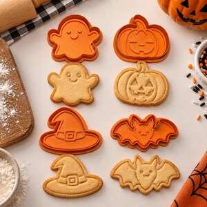 May include: A set of orange plastic cookie cutters and matching baked sugar cookies in the shapes of a ghost, a jack-o'-lantern, a witch hat, and a bat. The cutters and cookies are arranged in pairs on a light surface with flour and sprinkles.